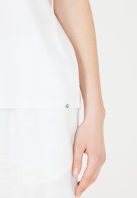 Valkoinen puuvillapaita, jossa on löysä malli, suora helma ja pieni brändilogo-tagi vasemmassa alareunassa. Sujuva tekstuuri.