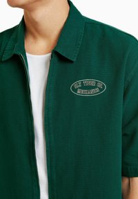 Chaqueta verde de cremallera y manga corta hecha de tela, con un parche de logo bordado que dice "Sly Tiger Co. Mechanics". Acabado texturizado.