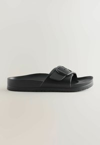 Schwarze Slip-On-Sandale mit dicker Sohle und verstellbarem Riemen mit großer abgerundeter Schnalle, auf einfarbig hellem Hintergrund.