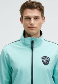 Giovane con capelli castano chiaro che indossa una giacca azzurra con zip e colletto alto, con un patch "Camp David" sul petto.