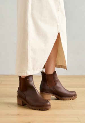 Brown ankle boots with a chunky heel, made of smooth leather with elastic side panels and textured rubber sole, paired with a beige skirt.