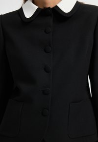 Svart figursydd kavaj med vit rund krage, täckta svarta knappar fram och två framfickor.