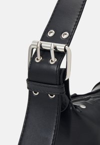 Sangle de sac en cuir noir avec boucle argentée à double ardillon et œillets en métal, attachée à un sac en cuir noir lisse avec tirette de fermeture éclair visible.