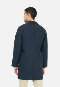 Cappotto blu navy con un design semplice e sartoriale, dotato di una fessura posteriore e un dettaglio con logo rosso sul colletto. Tessuto leggero, lunghezza fino a metà coscia.
