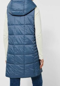 Fodrad blå väst med huva, med ett kviltat mönster och längre snitt. Mjuk, glänsande tyg med vertikala sömmar och inga synliga knäppningar.