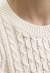 Nahaufnahme des krämfarbenen Kabelstrickpullovers am Halsausschnitt und oberen Brustbereich mit geripptem Kragen und detailliertem Muster.