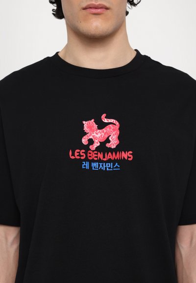 T-shirt noir en coton avec un graphisme léopard rouge et le texte "LES BENJAMINS" en rouge, accompagné d'un texte supplémentaire en bleu en dessous.