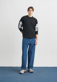 Μαύρη ριμπ φούτερ με άσπρη γραφική λεπτομέρεια στα μανίκια, συνδυασμένη με μπλε χαλαρής εφαρμογής τζιν και γκρι αθλητικά παπούτσια σε μπλε δάπεδο.