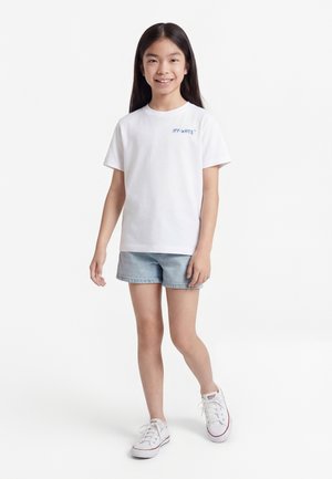 Lacht meisje met lang haar, een wit Off-White T-shirt, lichte denimshorts en witte sneakers, staand tegen een witte achtergrond.