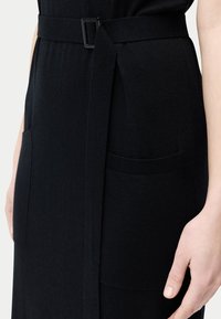 Svart klänning med texturerad stickning, sidofickor och ett midjebälte med rektangulärt spänne, som framhäver en figursydd siluett.