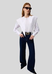 Donna che indossa una blusa bianca oversize con un ampio colletto ricamato, jeans larghi scuri, cintura nera, scarpe nere a punta e occhiali da sole tartarugati.
