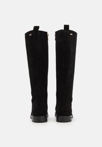 Bottes hautes en daim noir avec talons bas, fermetures éclair à l'arrière et petit logo drapeau près du sommet, présentées sur un fond blanc.