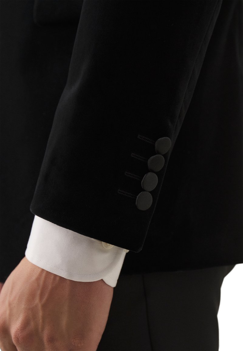Czarna aksamitna marynarka z rękawami, na których znajdują się cztery czarne guziki u mankietu, oraz biała koszula z zaokrąglonym mankietem widoczna poniżej.