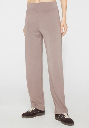 Personne portant un pantalon ample taupe et des baskets noires avec des lacets blancs, debout devant un fond clair uni.