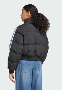 Jonge vrouw met krullend haar die iets is omgedraaid, gekleed in een zwarte pufferjack met witte strepen, gecombineerd met blauwe wijde jeans.