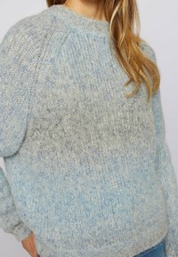 Een vrouw in een donzige gebreide trui in lichtblauwe en beige tinten, gecombineerd met blauwe jeans, gezien van schouders tot heupen.