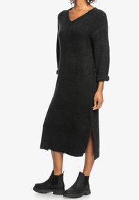 Schwarzes Strickkleid mit V-Ausschnitt, langen Ärmeln und seitlichen Schlitzen. Kombiniert mit schwarzen Stiefeletten mit elastischen Seiteneinsätzen.