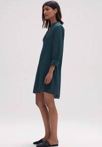 Langärmliges, knielanges Kleid aus dunkelblaugrünem, strukturiertem Stoff mit V-Ausschnitt und Seitenschlitzen, kombiniert mit schwarzen Slippern.