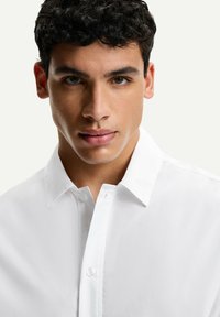 Camicia bianca con colletto classico e una leggera lucentezza. Tessuto liscio, design aderente, con un unico bottone al collo.