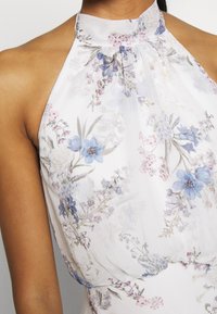 Närbild av en halterneck-klänning med vit bas och ljusblå, lila och rosa blommönster på slät tyg.