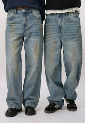 To personer iført løstsiddende, falmede blå jeans med sorte sko synlige, stående side om side mod en ensfarvet baggrund.