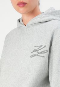 Donna che indossa un hoodie grigio chiaro con un piccolo logo ricamato in argento lucido sul petto, labbra chiuse e viso parzialmente visibile.
