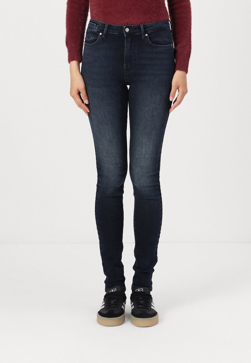 Blugi skinny de culoare albastru închis, din denim, cu cinci buzunare și închidere cu nasture și fermoar. Purtați cu adidași negri.