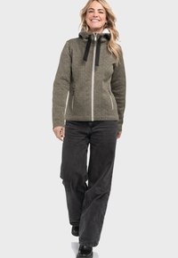 Eine lächelnde Frau, die mit einem braunen Reißverschlussjackett mit schwarzen Kordeln, schwarzen weit geschnittenen Hosen und schwarzen Schuhen auf einem weißen Hintergrund nach vorne geht.