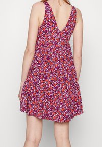 Květinové šaty s výstřihem do V, s fialovým základem zdobeným červenými, oranžovými a bílými květy; vrstvený, rozšířený lem.