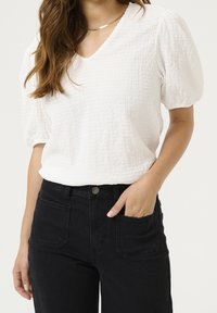 Weißes strukturiertes Oberteil mit Puffärmeln und V-Ausschnitt. Kombiniert mit hochtaillierten schwarzen Jeans mit Taschen und einem Metallknopfverschluss.