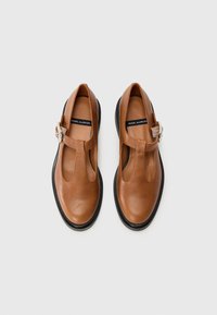 Chaussures T-bar en cuir marron avec un bout arrondi, semelles en caoutchouc noir et une sangle réglable. Texture lisse avec un design minimaliste.