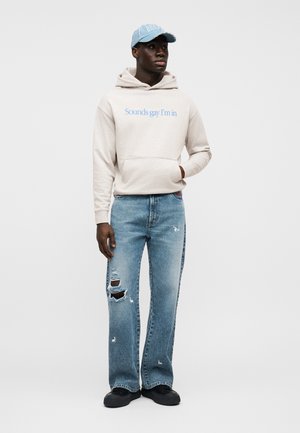 Beige hættetrøje med blå tekst, frontlomme; slidte lyseblå jeans med revner; sorte sko; lyseblå kasket.