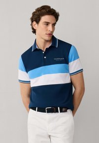 Joven con camisa tipo polo a rayas en azul marino, azul y blanco, pantalones blancos y un cinturón tejido azul marino, posando frente a un fondo liso.