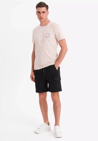Lichtbeige T-shirt met een gespikkeld patroon, rond embleem, gecombineerd met zwarte cargoshorts en witte sneakers. Model staat in een neutrale houding.
