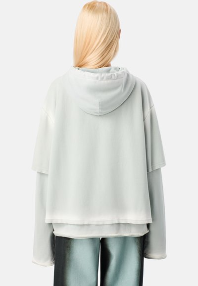 Jasnoszara, oversize bluza z kapturem o podwójnej warstwie, charakteryzująca się miękką teksturą. Ubranie ma krótkie rękawy i luźny krój.