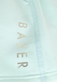 Ljust mintgrön tyg med en slät textur har ordet "BAKER" i metallisk guldtryck. Sidansömmen har en tryckt varumärkesetikett.