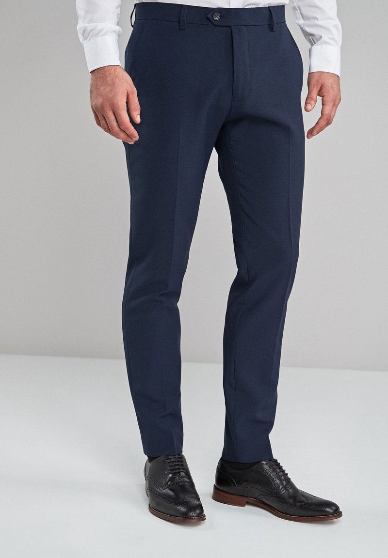 Mężczyzna ubrany w granatowe, dopasowane spodnie garniturowe, białą koszulę i czarne skórzane eleganckie buty stojący na jednolitym tle.