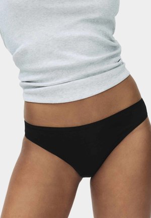 Frauenoberkörper, die ein weißes, geripptes Trägershirt und schwarze, tief sitzende Bikini-Unterwäsche vor einfarbigem Hintergrund trägt.