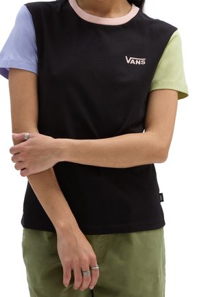 Personne portant un t-shirt Vans noir avec des manches violet clair et vert clair, un pantalon vert olive, bras croisés et bagues aux doigts.