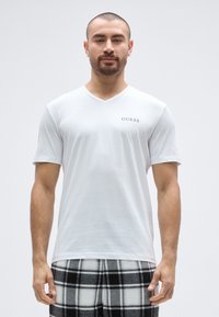Hvid V-hals t-shirt med et lille gråt "GUESS"-logo på venstre bryst. Blødt stof, afslappet pasform. Kombineret med sort-hvide ternede bukser.