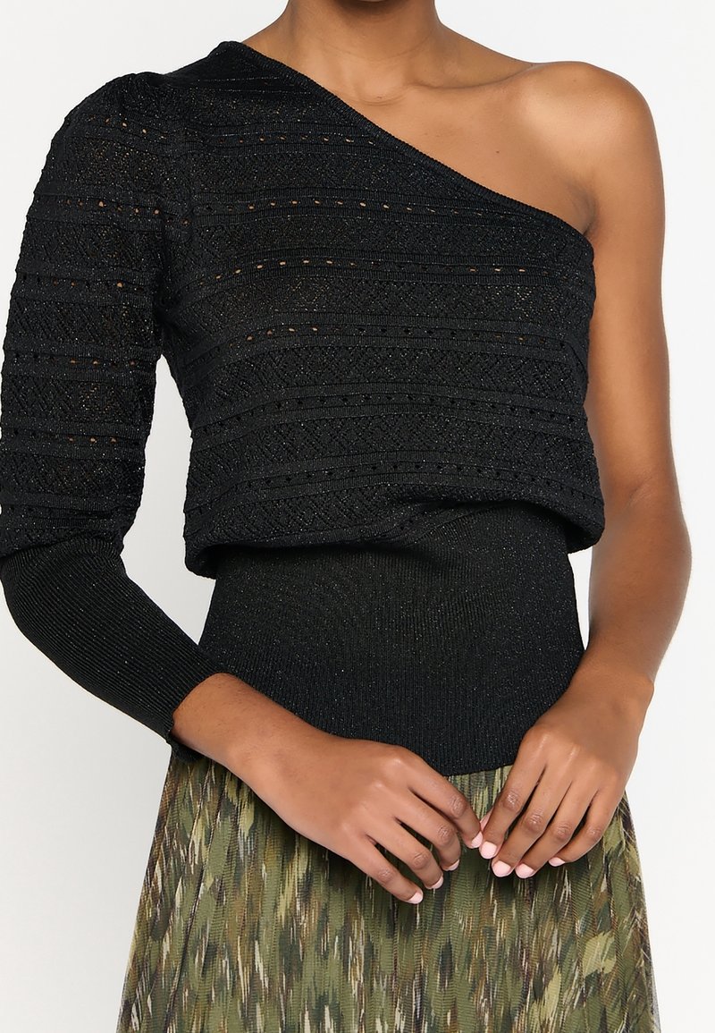 Femme portant un pull en maille noir à une épaule avec une manche longue et une jupe verte à motifs, les mains jointes devant.