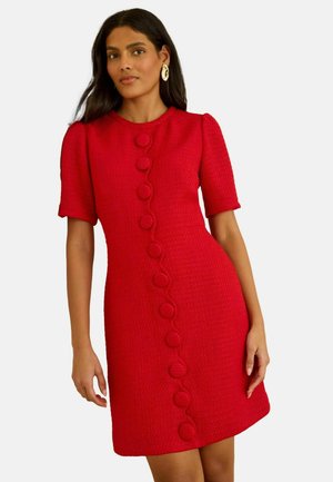 Nő texturált piros, rövid ujjú ruhában, amelyen nagy, textillel bevont gombok futnak végig az elején, arany karika fülbevalóval.