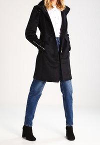 Cappotto nero su misura con cappuccio, tasche con zip e finitura opaca. Abbinato a jeans blu e stivaletti neri.