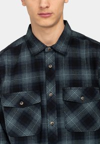 Flanellen shirt in donkerblauw en zwart geruit patroon. Voorzien van een button-down kraag en twee borstzakken met knoopsluitingen.
