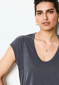 Mørkegrå T-shirt med korte ærmer, bred halsudskæring og blød tekstur. Har en diskret lomme og lag-på-lag look, parret med en delikat halskæde.