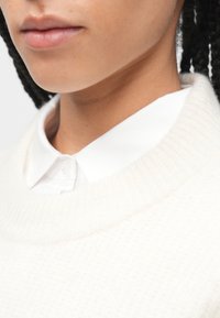 Valkoinen, tekstuuroitu neule, jossa on resorikaulus, yhdistettynä ryhdikkääseen valkoiseen napitettuun paitaan alla. Lähikuva pääntiestä ja kauluksesta.