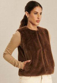 Brązowa kamizelka z sztucznego futra z okrągłym dekoltem, dwoma bocznymi kieszeniami i zapięciem na zatrzask z przodu, nałożona na beżową bluzkę z długim rękawem.