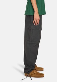 Cargo broek in donkergrauwe stof met een losse pasvorm, voorzien van een zijzak en omgeslagen hemden. Gecombineerd met bruine mocassinschoenen.