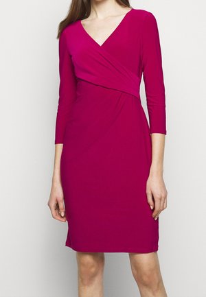 Nainen pukeutuneena istuvaan, polven alle ulottuvaan magentanväriseen kietaisumekkoon, jossa on kolmevartiset hihat ja V-pääntie, seisoo yksiväristä taustaa vasten.