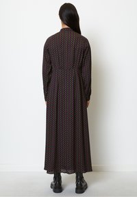 Robe noire à manches longues avec un motif géométrique répétitif rouge et vert, cintrée à la taille et atteignant les chevilles, accompagnée de bottes noires.
